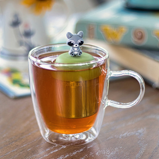 grey raccoon tea infuser for loose leaf tea with a fine mesh stainless steel tea strainer 
that reads FLOAT2INFUSE on a green base floating in a glass tea mug 
with books in the background