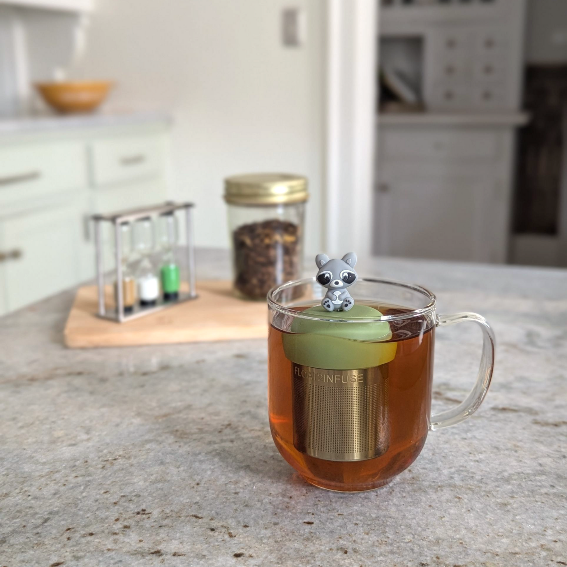 grey raccoon tea infuser for loose leaf tea  with a fine mesh stainless steel strainer that reads FLOAT2INFUSE on a green base floating in a glass tea mug with loose leaf tea in the background