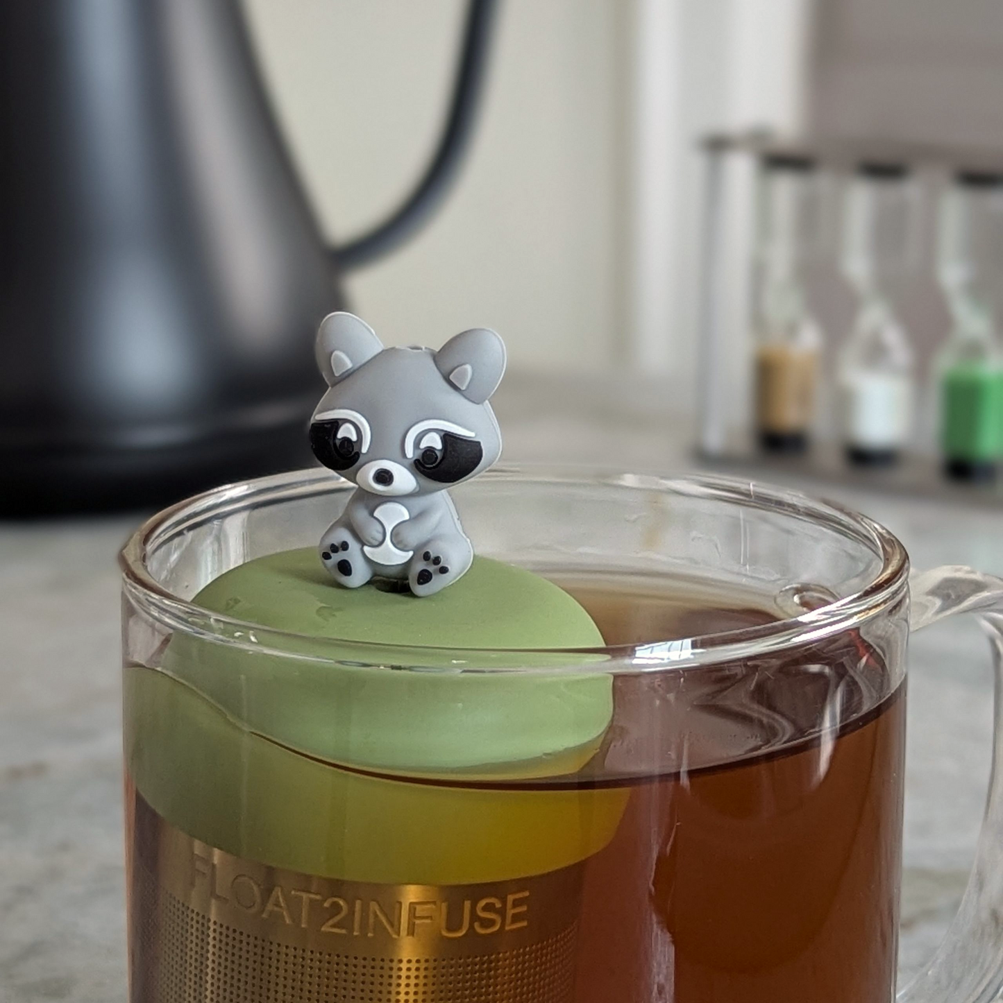 grey raccoon tea infuser for loose leaf tea  with a fine mesh stainless steel strainer that reads FLOAT2INFUSE on a green base floating in a glass tea mug with a tea kettle in the background