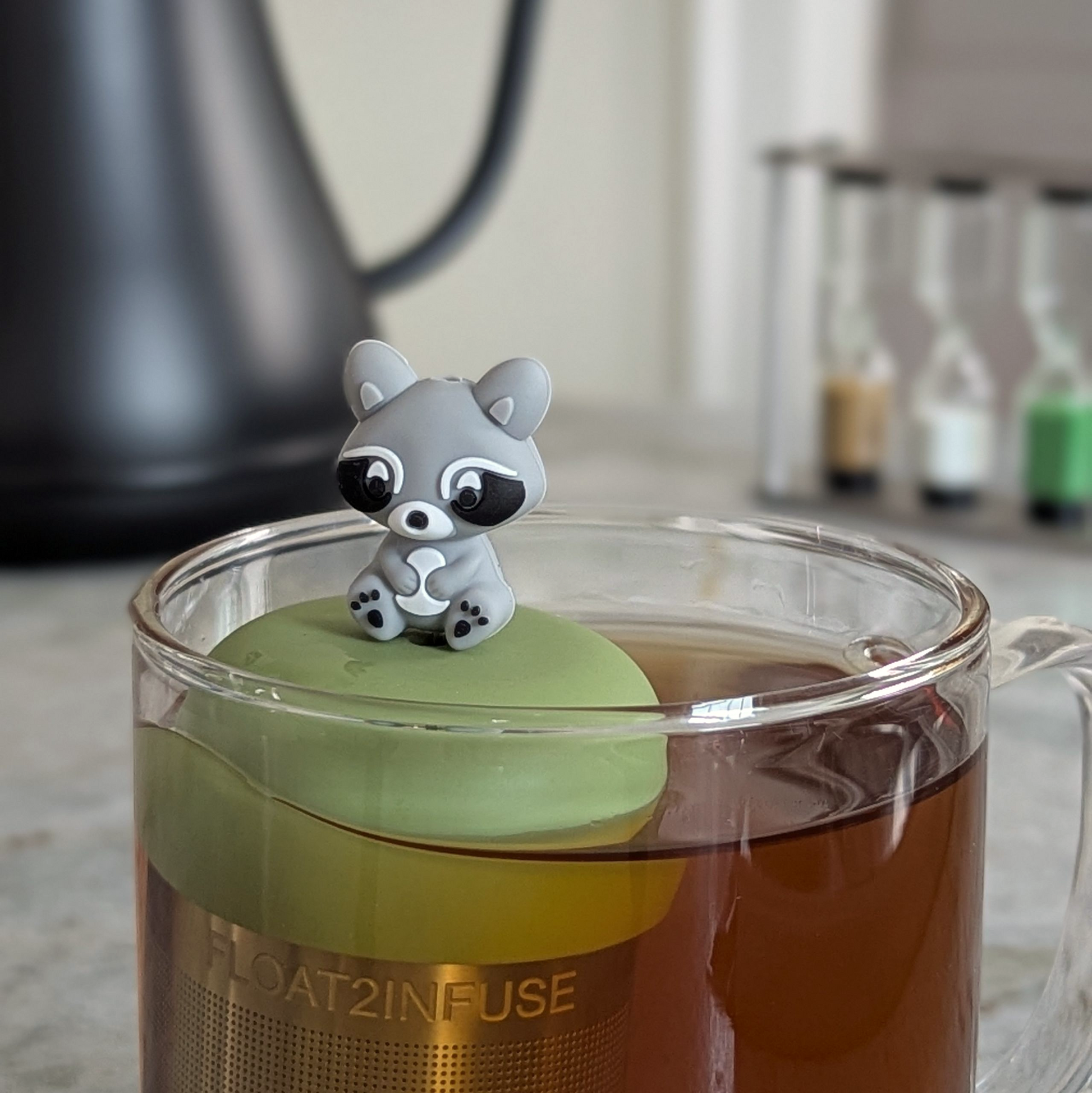 grey raccoon tea infuser for loose leaf tea  with a fine mesh stainless steel strainer that reads FLOAT2INFUSE on a green base floating in a glass tea mug with a tea kettle in the background