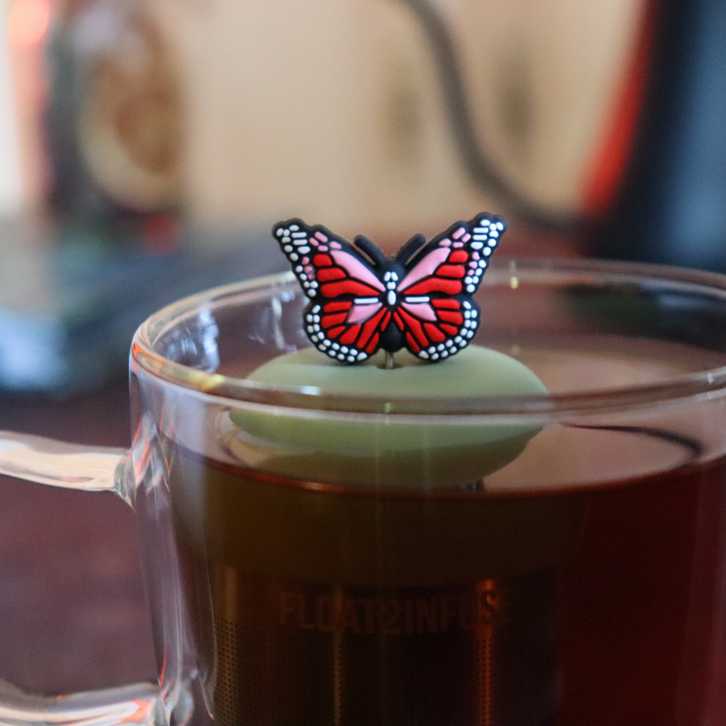 red and pink butterfly tea infuser for loose leaf tea  with a fine mesh stainless steel tea strainer that reads FLOAT2INFUSE on a green base floating in a glass tea mug with tea kettle in the background