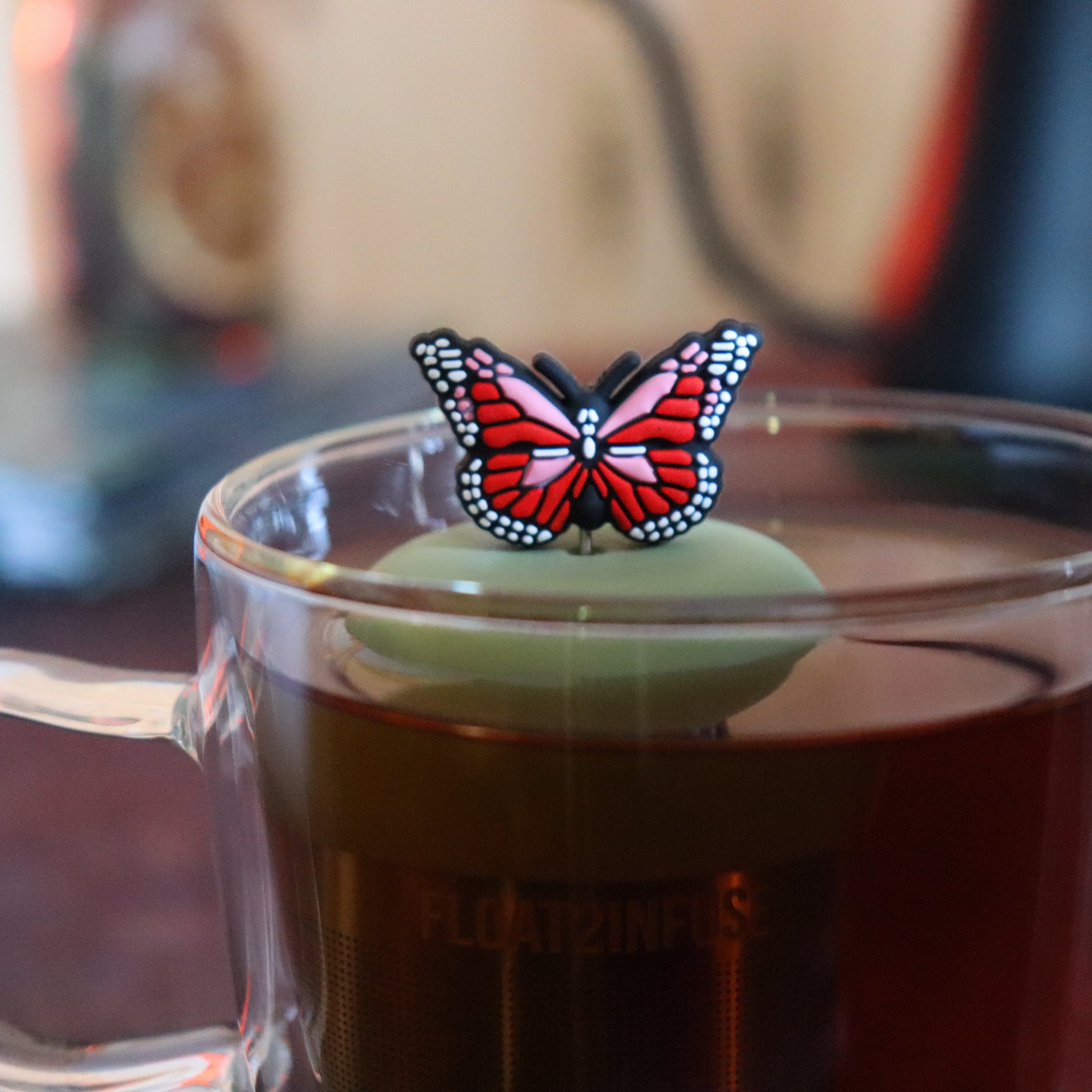 red and pink butterfly tea infuser for loose leaf tea  with a fine mesh stainless steel tea strainer that reads FLOAT2INFUSE on a green base floating in a glass tea mug with tea kettle in the background