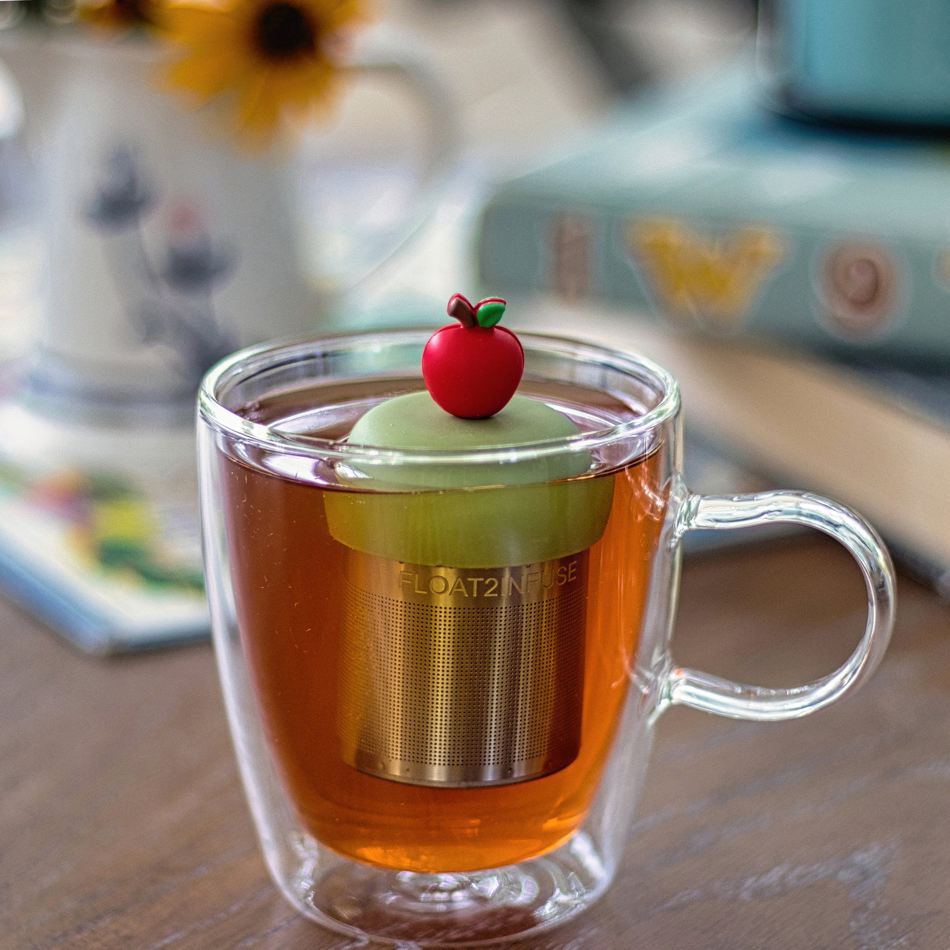 red apple tea infuser for loose leaf tea with a fine mesh stainless steel tea strainer that reads FLOAT2INFUSE on a green base  floating in a glass tea mug with books in the background