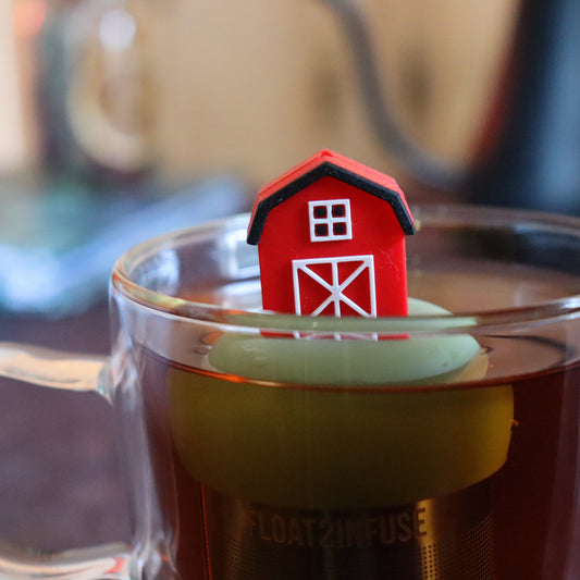 red barn tea infuser for loose leaf tea  with a fine mesh stainless steel tea strainer that reads FLOAT2INFUSE on a green base floating in a glass tea mug with tea kettle in the background