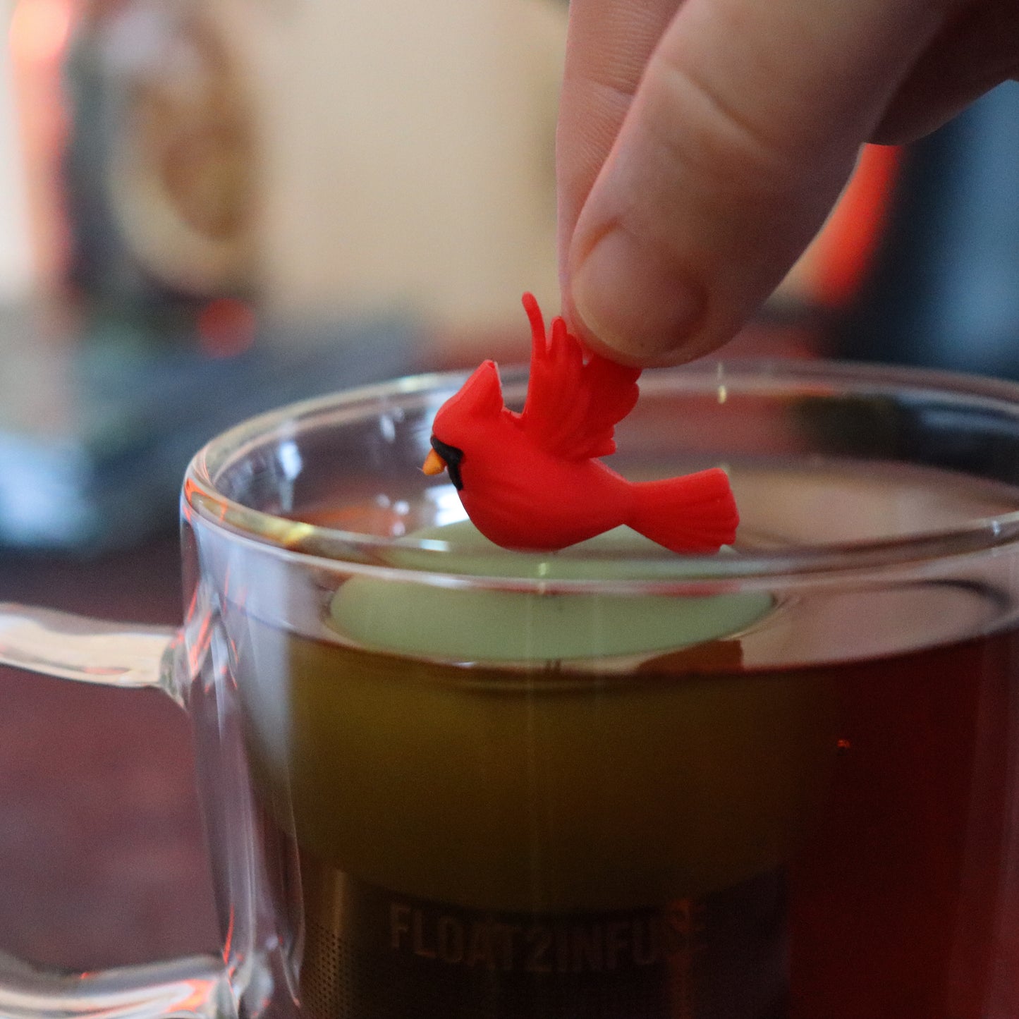 red cardinal bird animal tea infuser for loose leaf tea  with a fine mesh stainless steel tea strainer that reads FLOAT2INFUSE on a green base floating in a glass tea mug with tea kettle in the background