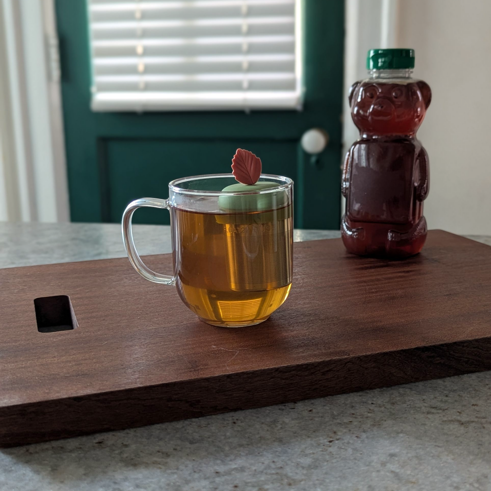 red leaf tea infuser for loose leaf tea  with a fine mesh stainless steel tea infuser that reads FLOAT2INFUSE on a green base floating in a glass tea mug with honey bear in the background