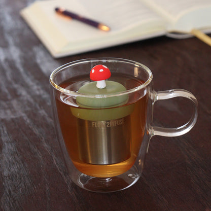 red mushroom tea infuser for loose leaf tea on a green base with a fine mesh stainless steel tea infuser that reads FLOAT2INFUSE floating in a glass tea mug with notebook and pen in the background