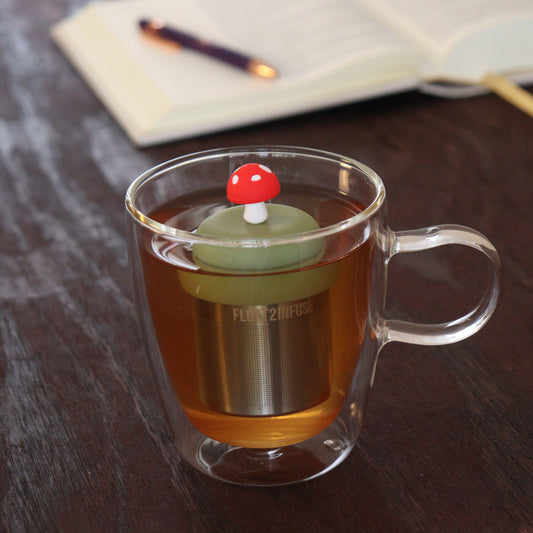 red mushroom tea infuser for loose leaf tea on a green base with a fine mesh stainless steel tea infuser that reads FLOAT2INFUSE floating in a glass tea mug with notebook and pen in the background