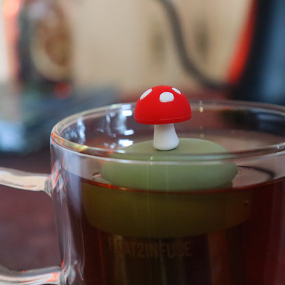 red mushroom tea infuser for loose leaf tea on a green base with a fine mesh stainless steel tea strainer that reads FLOAT2INFUSE floating in a glass tea mug with tea kettle in the background