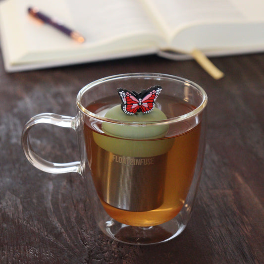red and pink butterfly tea infuser for loose leaf tea  with a fine mesh stainless steel tea infuser that reads FLOAT2INFUSE on a green base floating in a glass tea mug with notebook and pen in the background