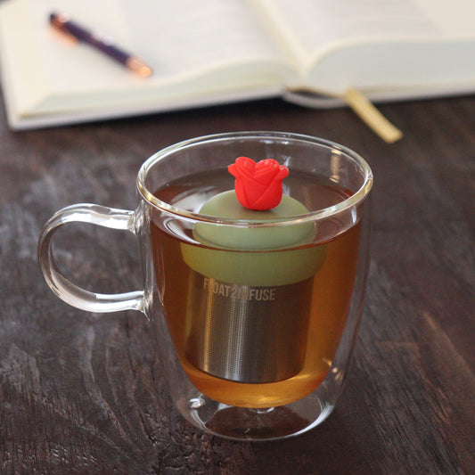 red rose animal tea infuser for loose leaf tea on a green base with a fine mesh stainless steel tea infuser that reads FLOAT2INFUSE floating in a glass tea mug with notebook and pen in the background