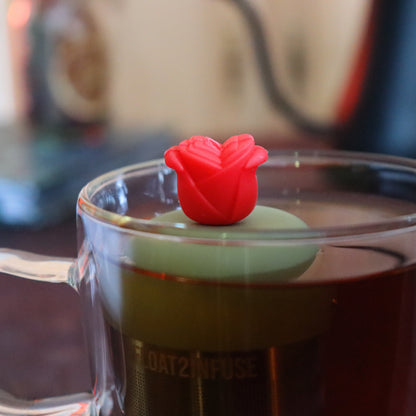 red rose tea infuser for loose leaf tea  with a fine mesh stainless steel tea strainer that reads FLOAT2INFUSE on a green base floating in a glass tea mug with tea kettle in the background