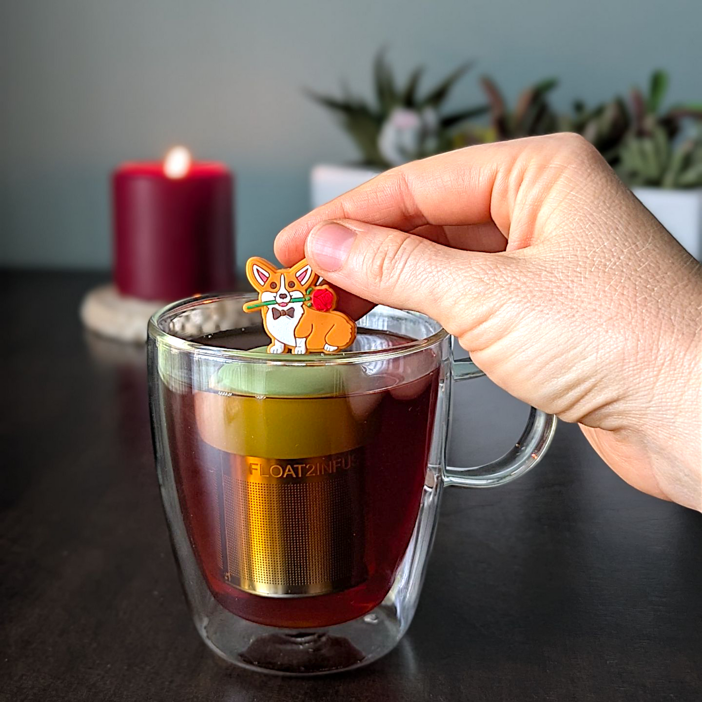 Orange and white corgi dog wearing a bowtie and holding a red rose in his mouth  tea infuser for loose leaf tea with a fine mesh stainless steel tea strainer that reads FLOAT2INFUSE on a green base with a lit candle and a plant in the background
