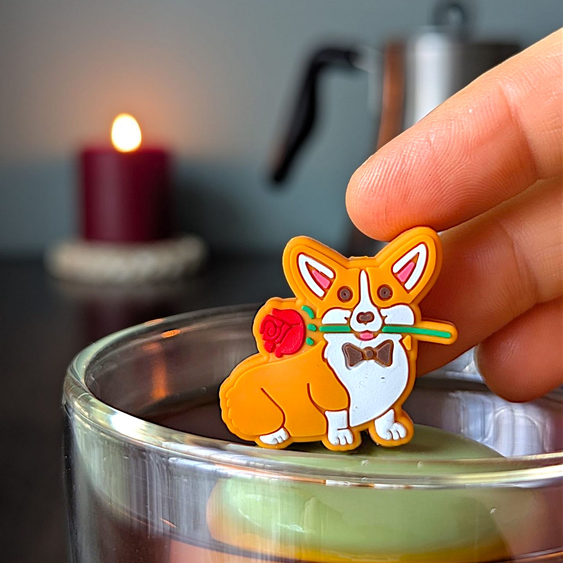 Orange and white corgi dog wearing a bowtie and holding a red rose in his mouth  tea infuser for loose leaf tea with a fine mesh stainless steel tea strainer that reads FLOAT2INFUSE on a green base with a lit candle and a tea kettle in the background