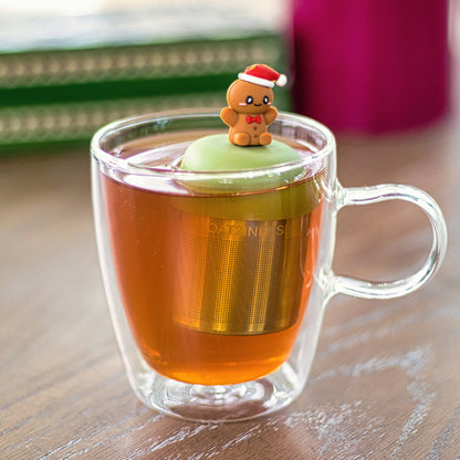 happy gingerbread man with a red bowtie and wearing a red and white santa hat   tea infuser for loose leaf tea with a fine mesh stainless steel tea strainer that reads FLOAT2INFUSE on a green base  floating in a glass tea mug 
with books in the background