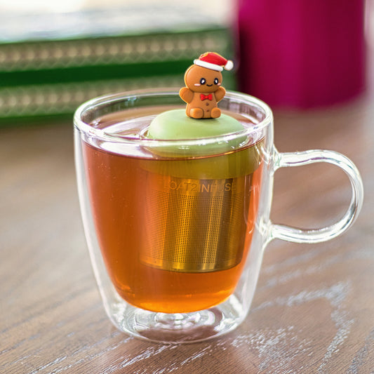 happy gingerbread man with a red bowtie and wearing a red and white santa hat   tea infuser for loose leaf tea with a fine mesh stainless steel tea strainer that reads FLOAT2INFUSE on a green base  floating in a glass tea mug 
with books in the background