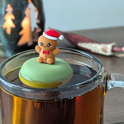 happy gingerbread man with a red bowtie and wearing a red and white santa hat   tea infuser for loose leaf tea with a fine mesh stainless steel tea strainer that reads FLOAT2INFUSE on a green base  floating in a glass tea mug 
with a candle in the background