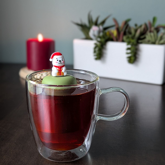 Smiling white polar bear holding a present and wearing a red santa hat tea infuser for loose leaf tea with a fine mesh stainless steel tea strainer that reads FLOAT2INFUSE on a green base  with a plants and a candle in the background