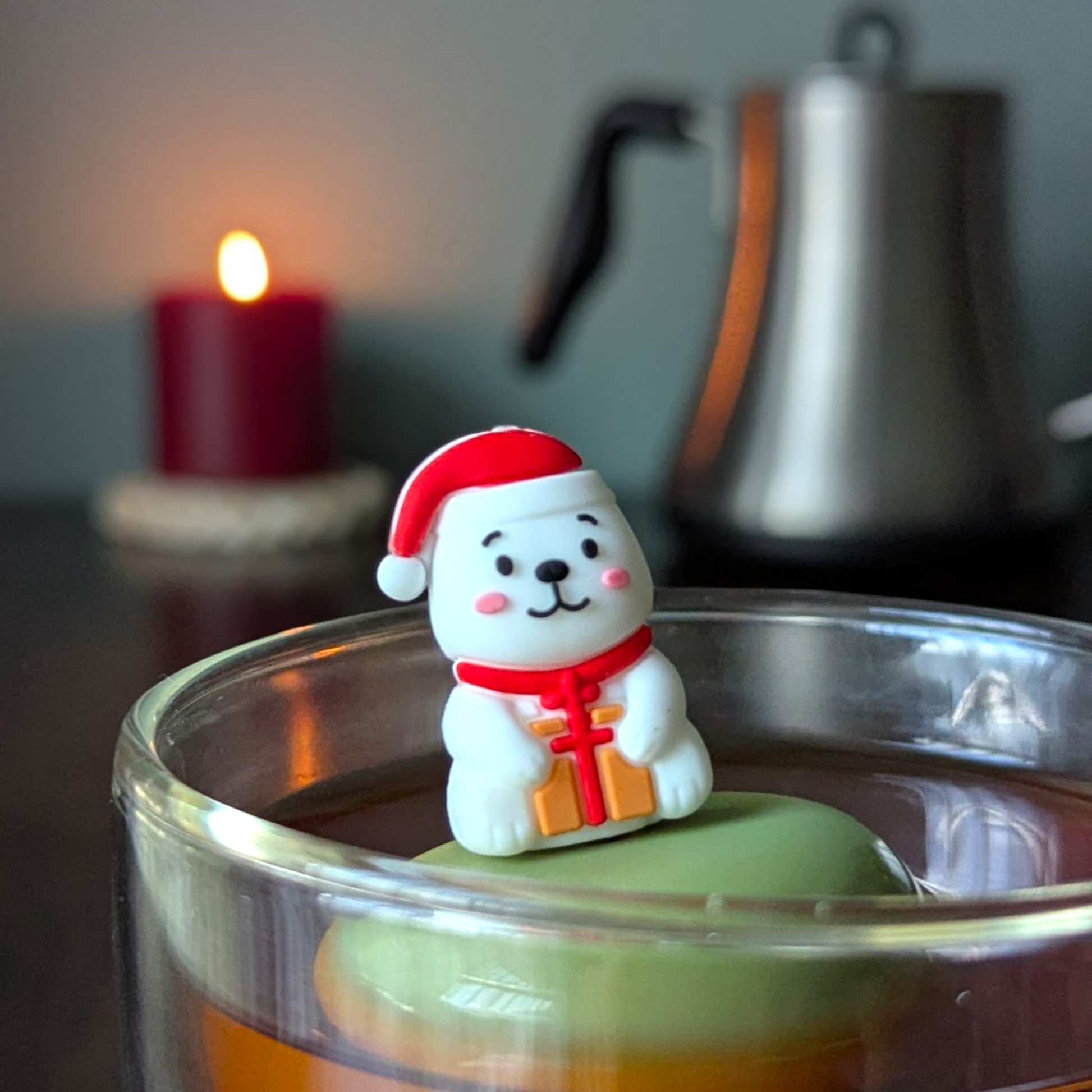 Smiling white polar bear holding a present and wearing a red santa hat tea infuser for loose leaf tea with a fine mesh stainless steel tea strainer that reads FLOAT2INFUSE on a green base  with a tea kettle and a candle in the background