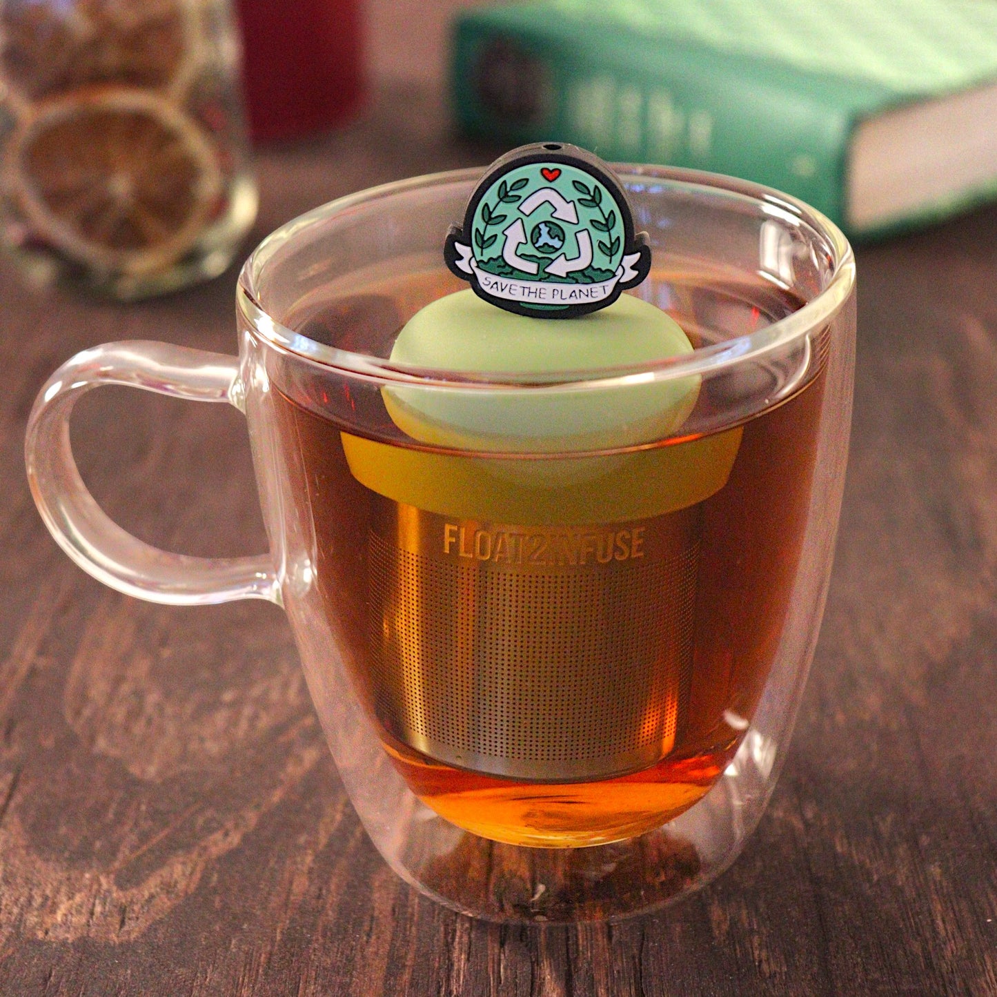  black circle with an image of the earth inside a recycling symbol, and text that reads "save the planet" for loose leaf tea with a fine mesh stainless steel tea infuser that reads FLOAT2INFUSE on a green base  floating in a glass tea mug with  loose leaf tea and sunglasses in the background