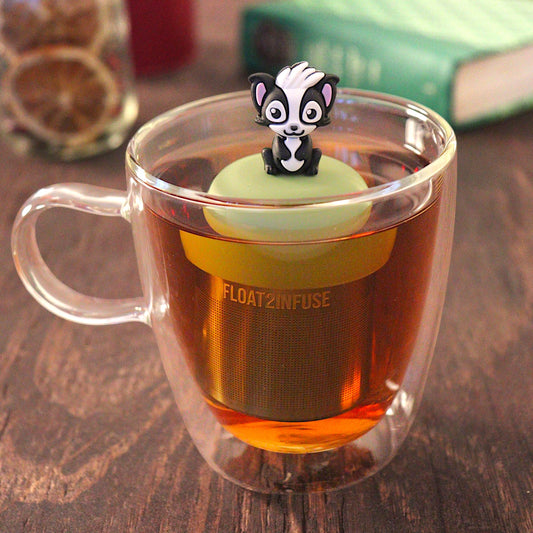 black and white skunk animal holding a red heart  tea infuser for loose leaf tea with a fine mesh stainless steel tea infuser that reads FLOAT2INFUSE on a green base  floating in a glass tea mug with  loose leaf tea and books in the background