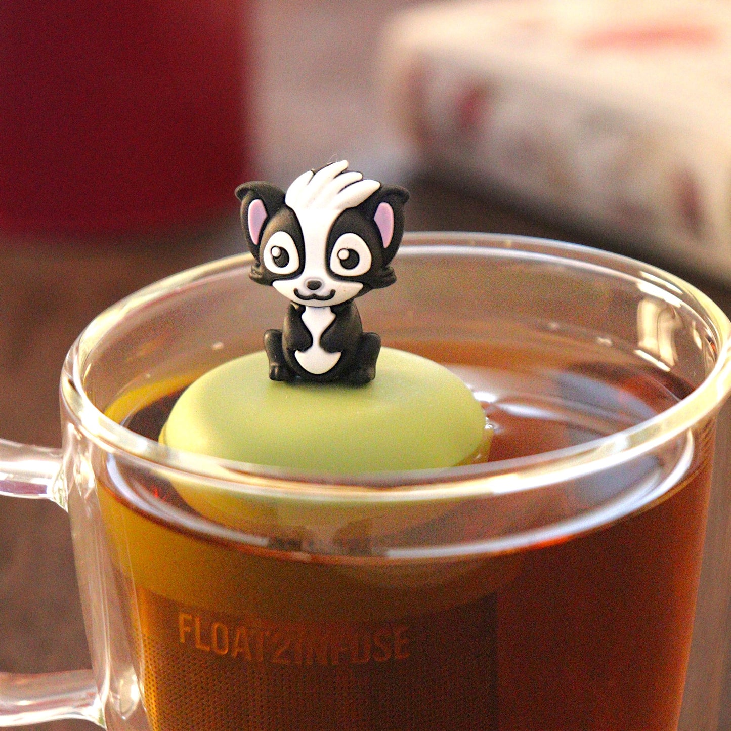 black and white skunk animal holding a red heart  tea infuser for loose leaf tea with a fine mesh stainless steel tea infuser that reads FLOAT2INFUSE on a green base  floating in a glass tea mug with  loose leaf tea and books in the background