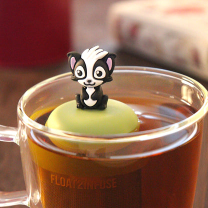 black and white skunk animal holding a red heart  tea infuser for loose leaf tea with a fine mesh stainless steel tea infuser that reads FLOAT2INFUSE on a green base  floating in a glass tea mug with  loose leaf tea and books in the background