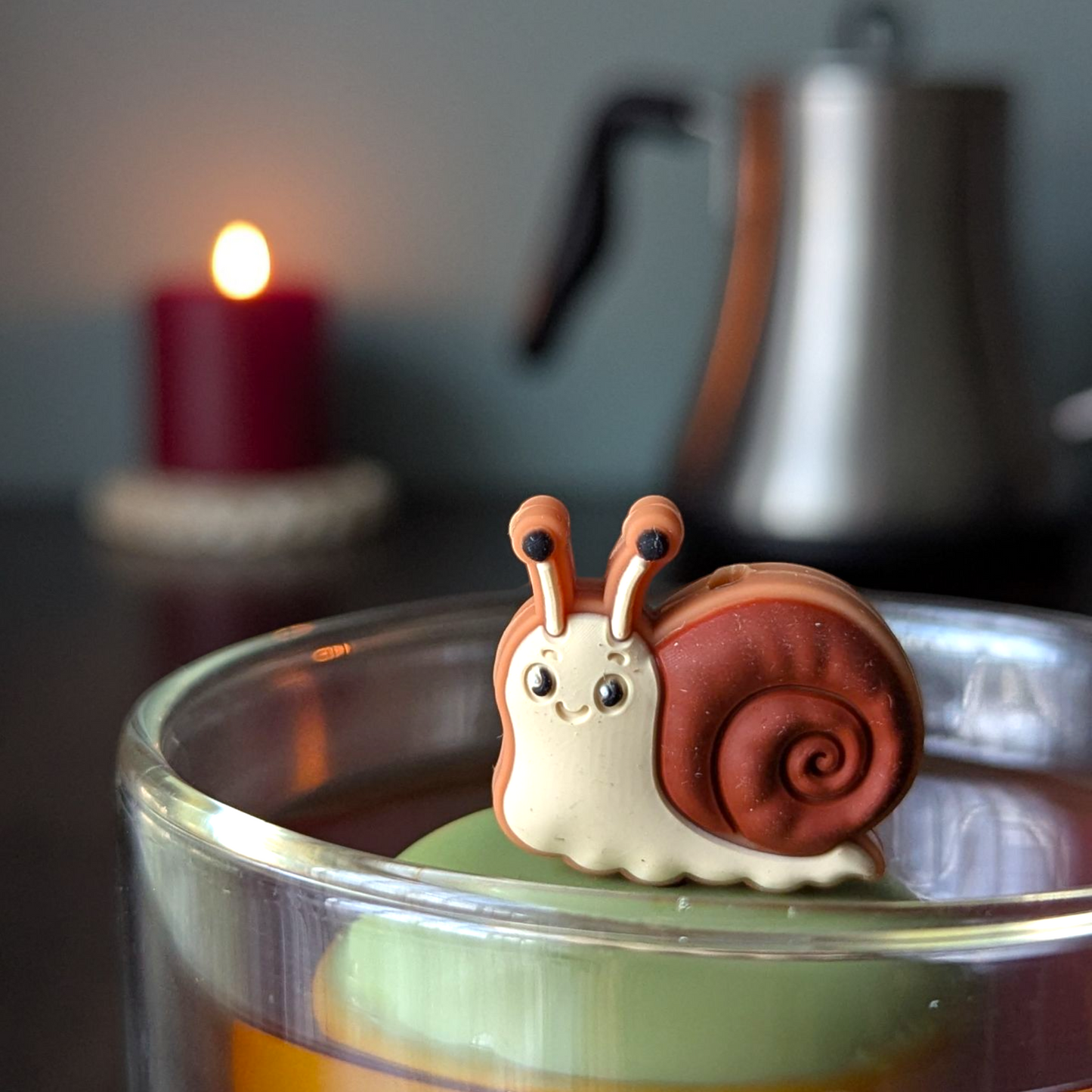 smiling brown snail tea infuser for loose leaf tea with a fine mesh stainless steel tea strainer that reads FLOAT2INFUSE on a green base with a lit candle and a tea kettle in the background