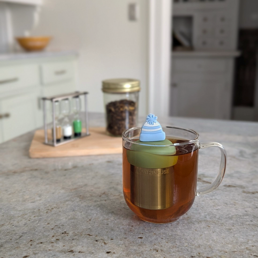 blue and white striped beanie hat tea infuser for loose leaf tea  with a fine mesh stainless steel strainer that reads FLOAT2INFUSE on a green base floating in a glass tea mug with loose leaf tea in the background