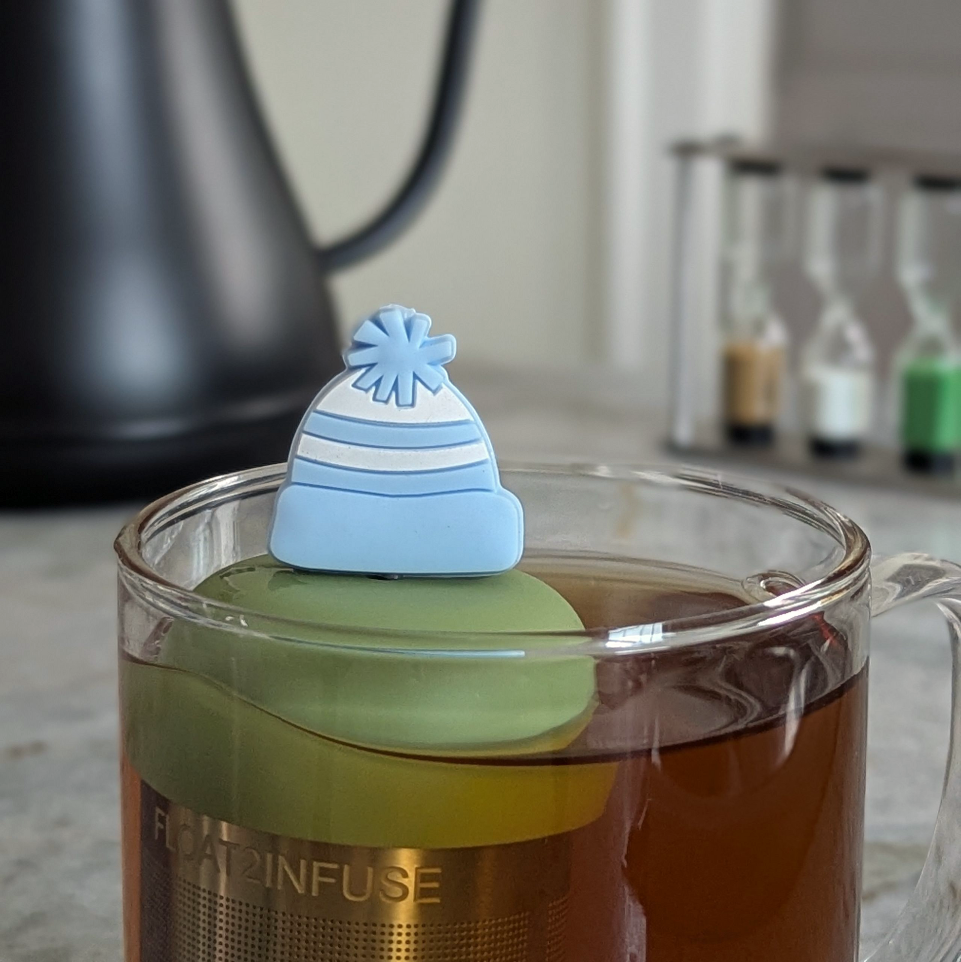blue and white striped beanie hat tea infuser for loose leaf tea  with a fine mesh stainless steel strainer that reads FLOAT2INFUSE on a green base floating in a glass tea mug with tea kettle in the background