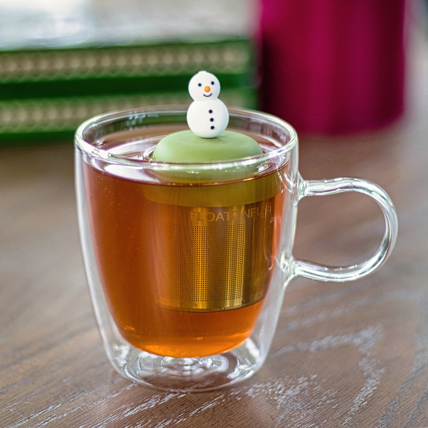smiling white snowman with orange carrot nose and black buttons tea infuser for loose leaf tea with a fine mesh stainless steel tea strainer that reads FLOAT2INFUSE on a green base  floating in a glass tea mug 
with books in the background