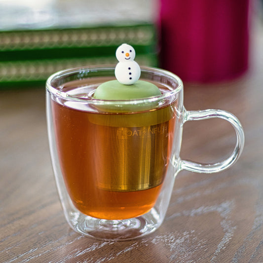smiling white snowman with orange carrot nose and black buttons tea infuser for loose leaf tea with a fine mesh stainless steel tea strainer that reads FLOAT2INFUSE on a green base  floating in a glass tea mug 
with books in the background