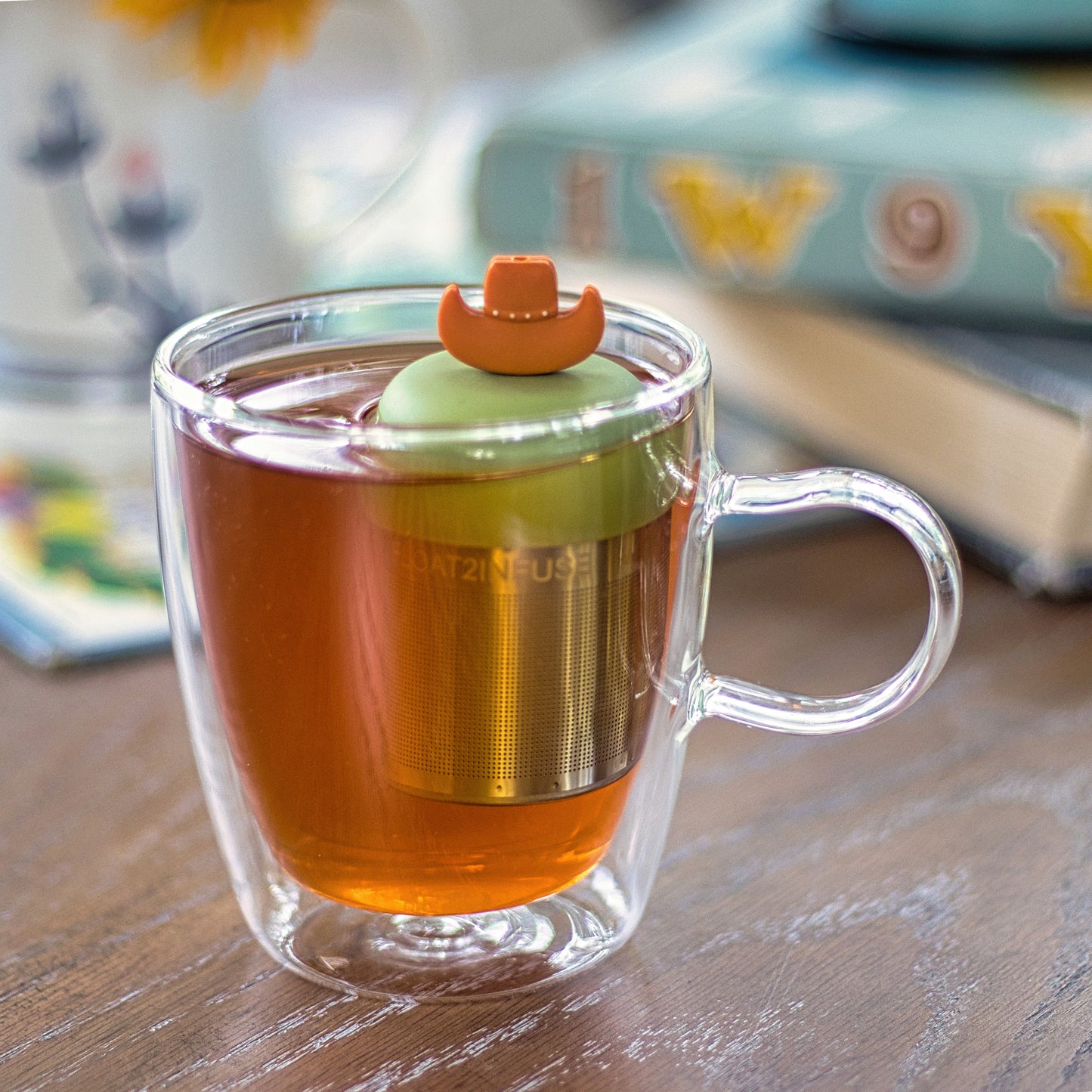 orange sombrero hat tea infuser for loose leaf tea with a fine mesh stainless steel tea strainer that reads FLOAT2INFUSE on a green base  floating in a glass tea mug 
with flowers in the background