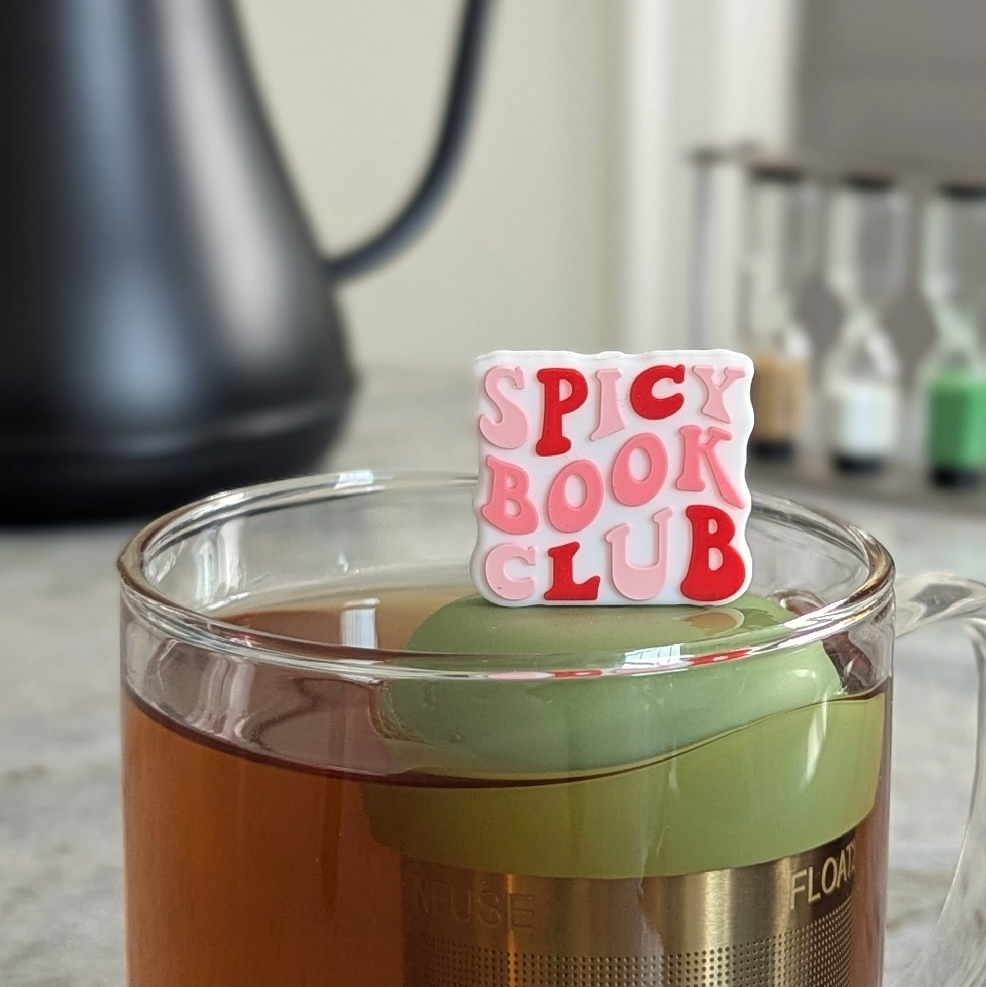 white square with text that reads "spicy book club" in pink and red letters tea infuser for loose leaf tea  with a fine mesh stainless steel strainer that reads FLOAT2INFUSE on a green base floating in a glass tea mug with tea kettle in the background