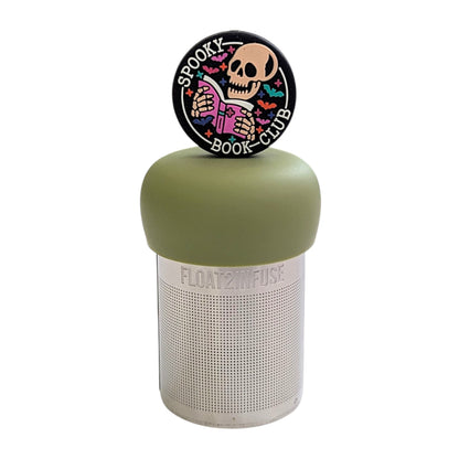 Black circle with an ivory skeleton reading a pink book with the text "spooky book club" and multicolored bats circling it tea infuser for loose leaf tea with a fine mesh stainless steel tea strainer that reads FLOAT2INFUSE on a green base with a white background