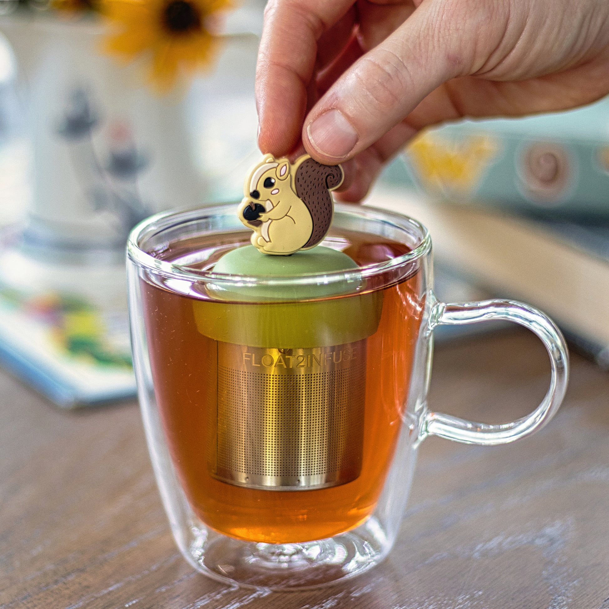 cute squirrel holding an acorn tea infuser for loose leaf tea with a fine mesh stainless steel tea strainer that reads FLOAT2INFUSE on a green base  floating in a glass tea mug 
with books in the background