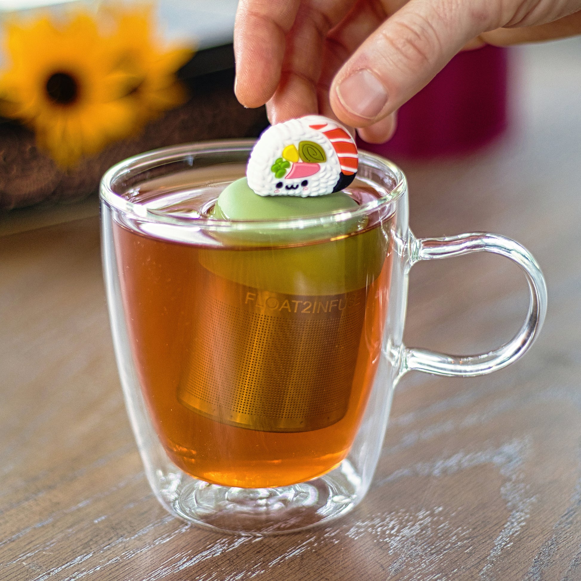 smiling face sushi tea infuser for loose leaf tea with a fine mesh stainless steel tea strainer that reads FLOAT2INFUSE on a green base  floating in a glass tea mug 
with flowers in the background