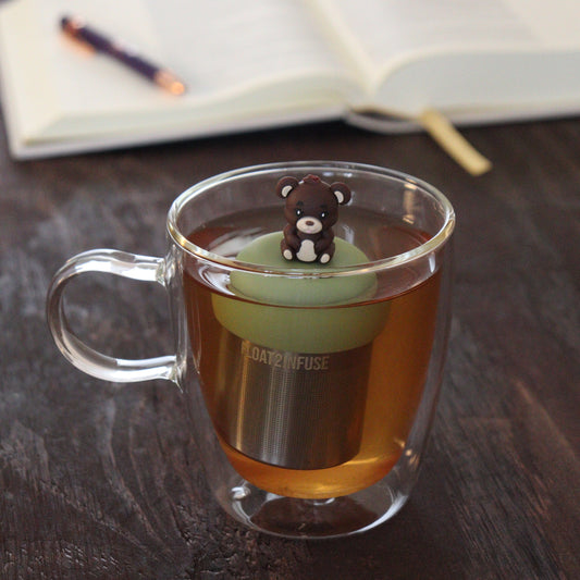 brown teddy bear sticking it's tongue out animal tea infuser for loose leaf tea with a fine mesh stainless steel tea infuser that reads FLOAT2INFUSE on a green base floating in a glass tea mug with notebook and pen in the background