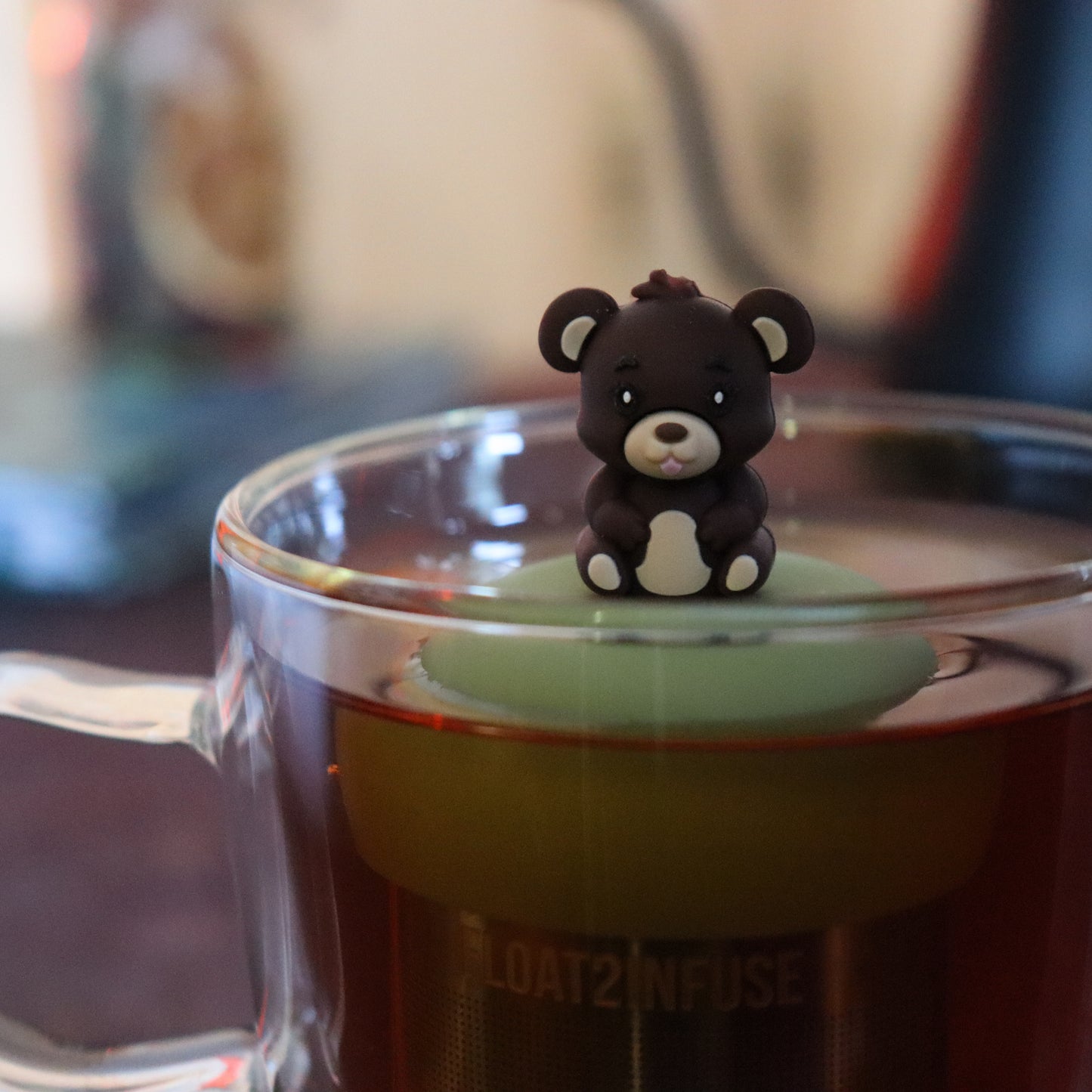 brown teddy bear animal sticking it's tongue out tea infuser for loose leaf tea  with a fine mesh stainless steel tea strainer that reads FLOAT2INFUSE on a green base floating in a glass tea mug with tea kettle in the background