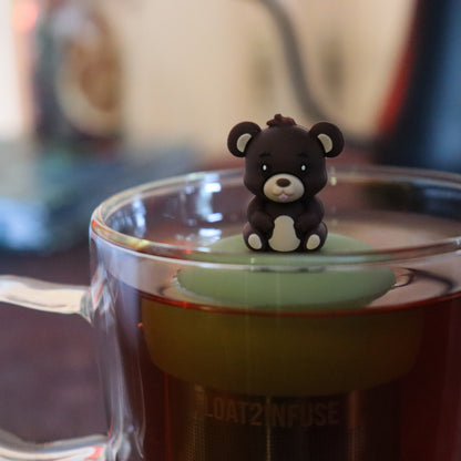 brown teddy bear animal sticking it's tongue out tea infuser for loose leaf tea  with a fine mesh stainless steel tea strainer that reads FLOAT2INFUSE on a green base floating in a glass tea mug with tea kettle in the background