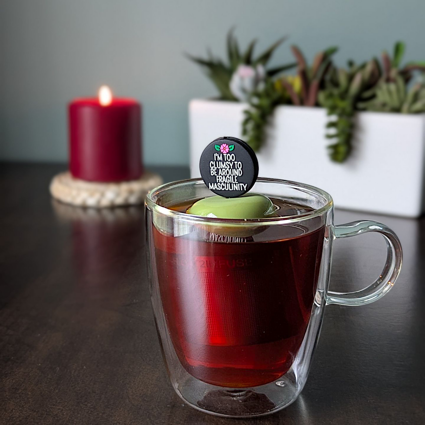 black circle with white text that reads "I'm too clumsy to be around fragile masculinity" with a pink  flower tea infuser  for loose leaf tea with fine mesh stainless steel tea strainer that reads FLOAT2INFUSE on a green base with a lit candle and plants in the background