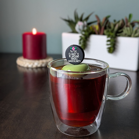 black circle with white text that reads "I'm too clumsy to be around fragile masculinity" with a pink  flower tea infuser  for loose leaf tea with fine mesh stainless steel tea strainer that reads FLOAT2INFUSE on a green base with a lit candle and plants in the background