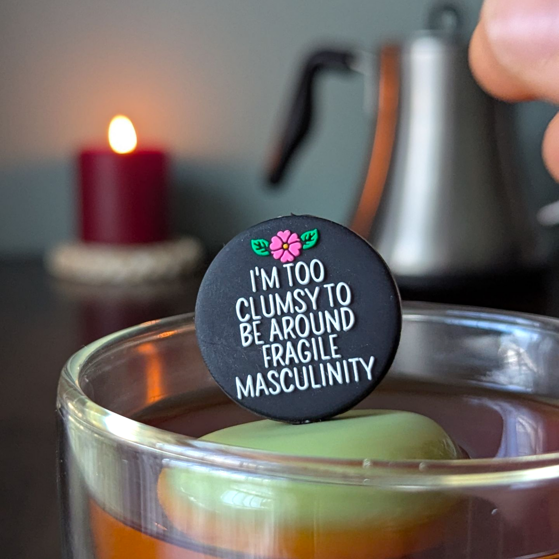 black circle with white text that reads "I'm too clumsy to be around fragile masculinity" with a pink  flower tea infuser  for loose leaf tea with fine mesh stainless steel tea strainer that reads FLOAT2INFUSE on a green base with a lit candle and a tea kettle in the background