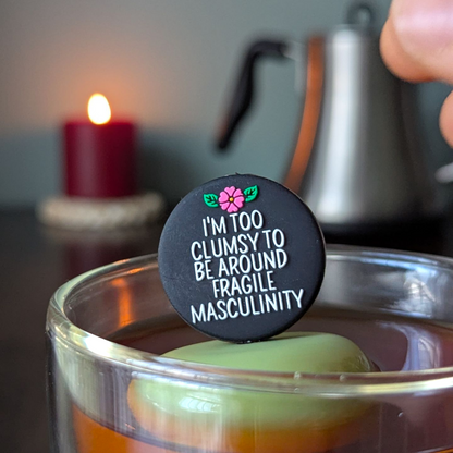 black circle with white text that reads "I'm too clumsy to be around fragile masculinity" with a pink  flower tea infuser  for loose leaf tea with fine mesh stainless steel tea strainer that reads FLOAT2INFUSE on a green base with a lit candle and a tea kettle in the background