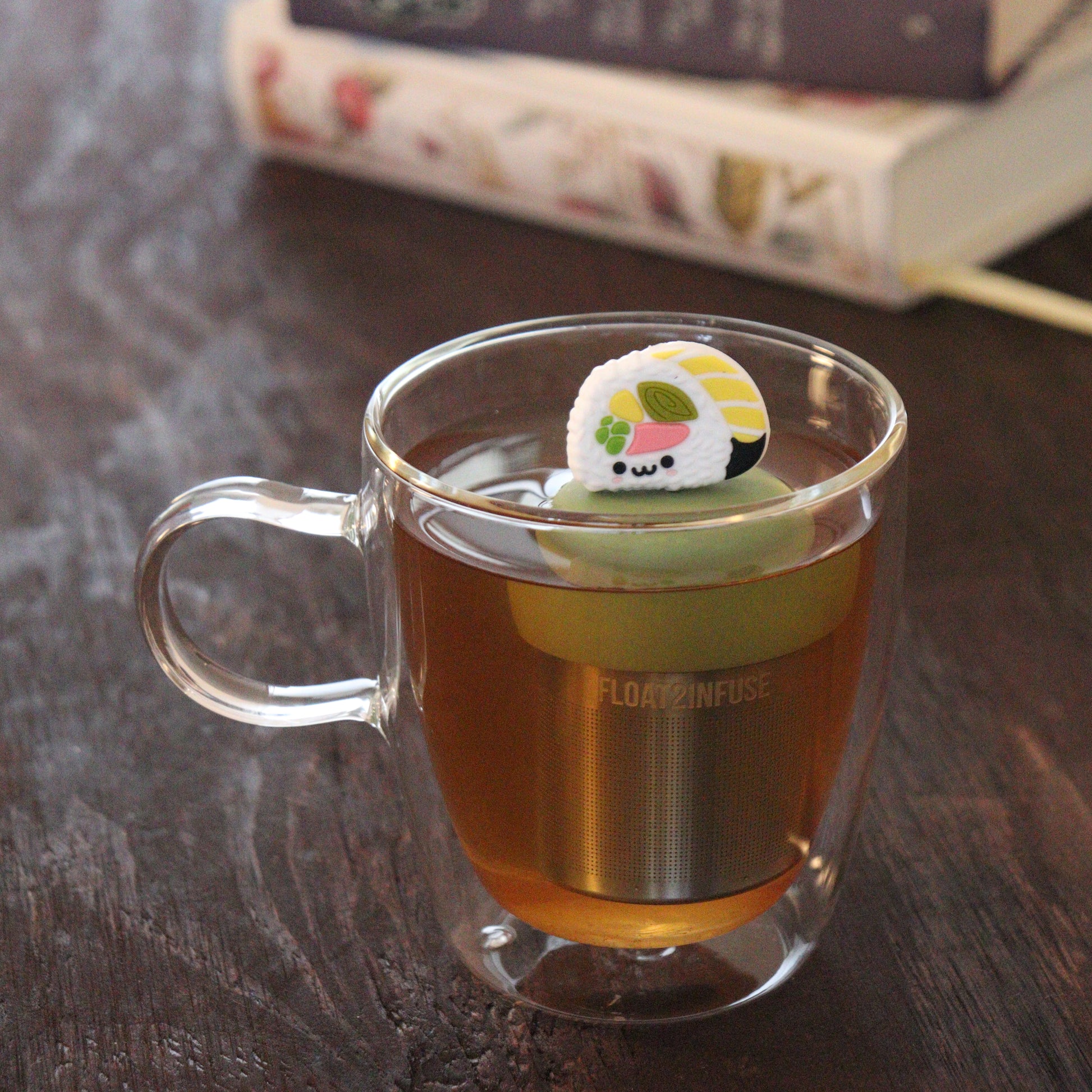 smiling white sushi tea infuser for loose leaf tea  with a fine mesh stainless steel tea infuser that reads FLOAT2INFUSE on a green base floating in a glass tea mug with books in the background