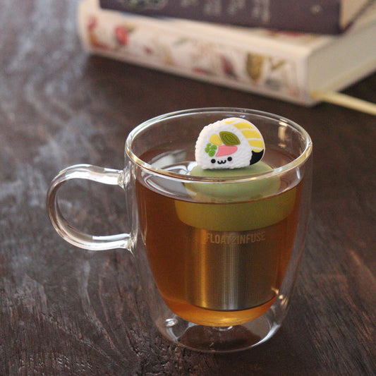 smiling white sushi tea infuser for loose leaf tea  with a fine mesh stainless steel tea infuser that reads FLOAT2INFUSE on a green base floating in a glass tea mug with books in the background