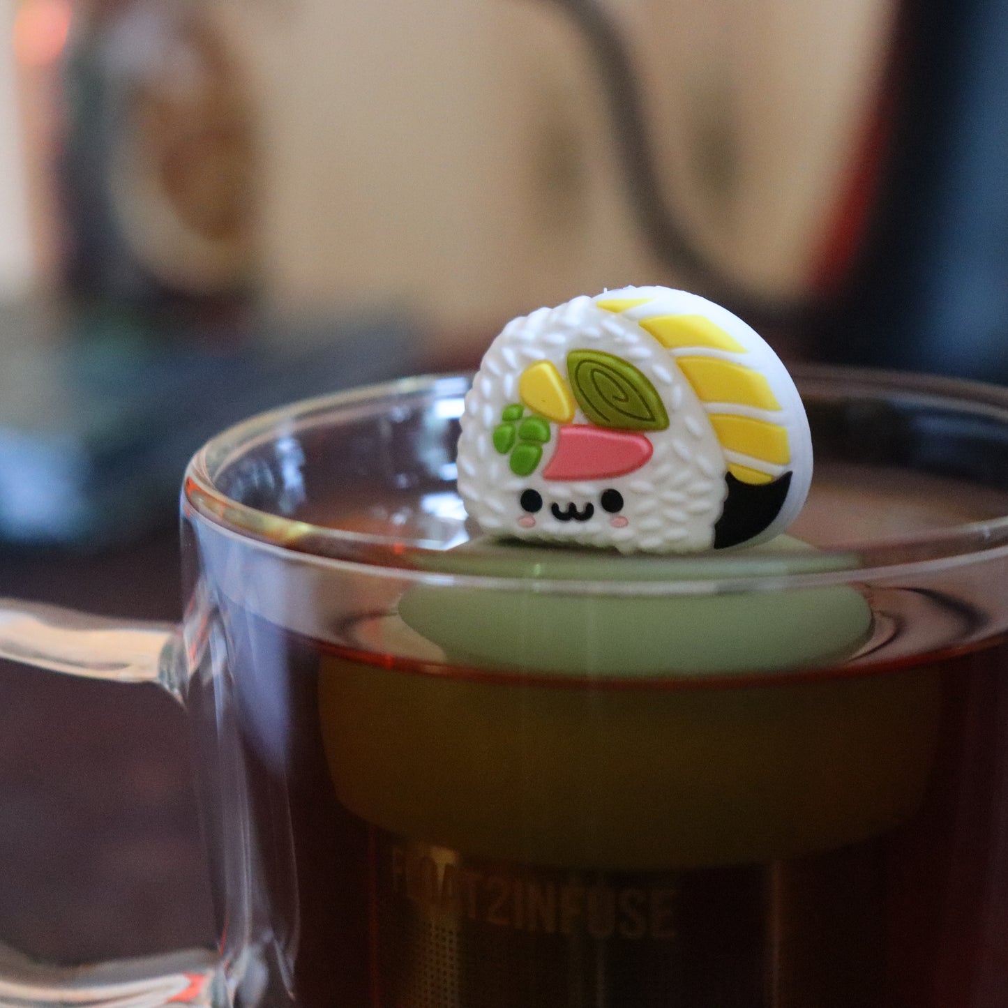 cute white sushi roll tea infuser for loose leaf tea  with a fine mesh stainless steel tea strainer that reads FLOAT2INFUSE on a green base floating in a glass tea mug with tea kettle in the background