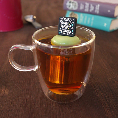 Black square with pink text that reads "very demure, very mindful, very cutesy" with pink and teal flowers and a pink bow tea infuser for loose leaf tea with a fine mesh stainless steel tea strainer that reads FLOAT2INFUSE on a green base with a tea canister and books in the background