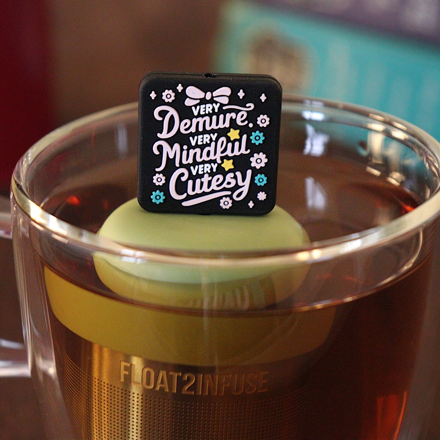 Black square with pink text that reads "very demure, very mindful, very cutesy" with pink and teal flowers and a pink bow tea infuser for loose leaf tea with a fine mesh stainless steel tea strainer that reads FLOAT2INFUSE on a green base with a tea canister and books in the background