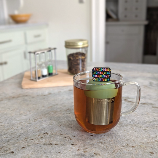 black rectangle with text "a well read woman is a dangerous creature" in rainbow font tea infuser for loose leaf tea  with a fine mesh stainless steel strainer that reads FLOAT2INFUSE on a green base floating in a glass tea mug with loose leaf tea in the background